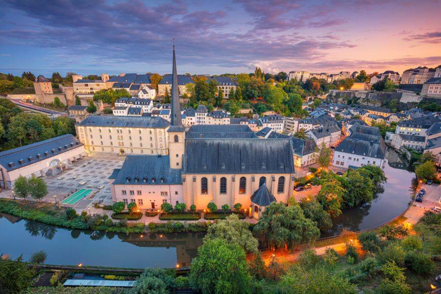 Blick auf die Abtei Neumünster und die Altstadt von Luxemburg-Stadt bei Sonnenuntergang, mit spiegelndem Fluss Alzette im Vordergrund.