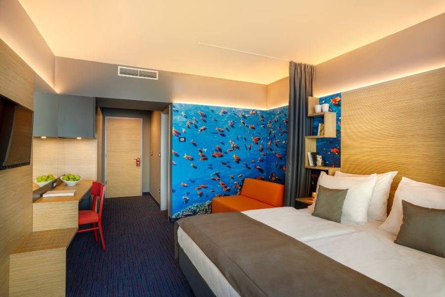 Ein farbenfrohes und gemütliches Hotelzimmer mit einer Meeres-Themenwand, auf der bunte Fische in einem blauen Unterwasserhintergrund dargestellt sind. Es gibt ein großes Doppelbett mit weißen Kissen und brauner Bettdecke.