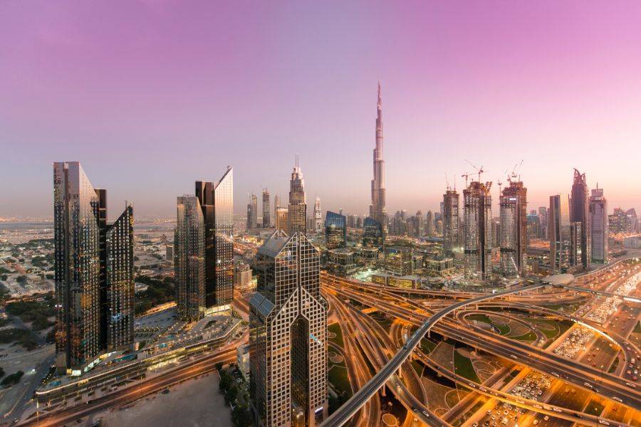 Panoramablick auf die moderne Skyline von Dubai mit dem Burj Khalifa im Zentrum, umgeben von Hochhäusern, Straßen und einem violett getönten Abendhimmel.