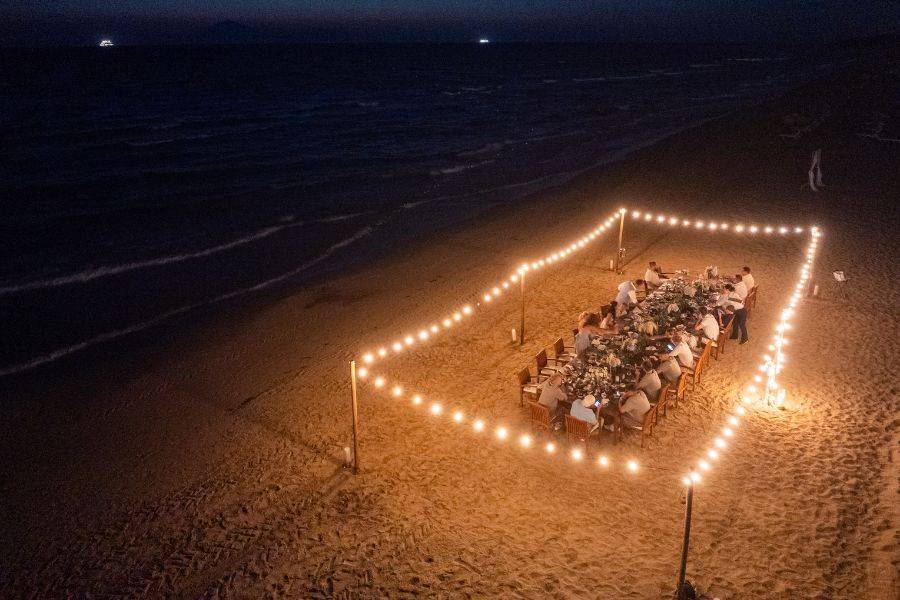 Festlich gedeckte Hochzeitstafel am Strand von Griechenland, umrahmt von Lichterketten, Gäste genießen das Dinner bei Nacht mit Meeresrauschen im Hintergrund.