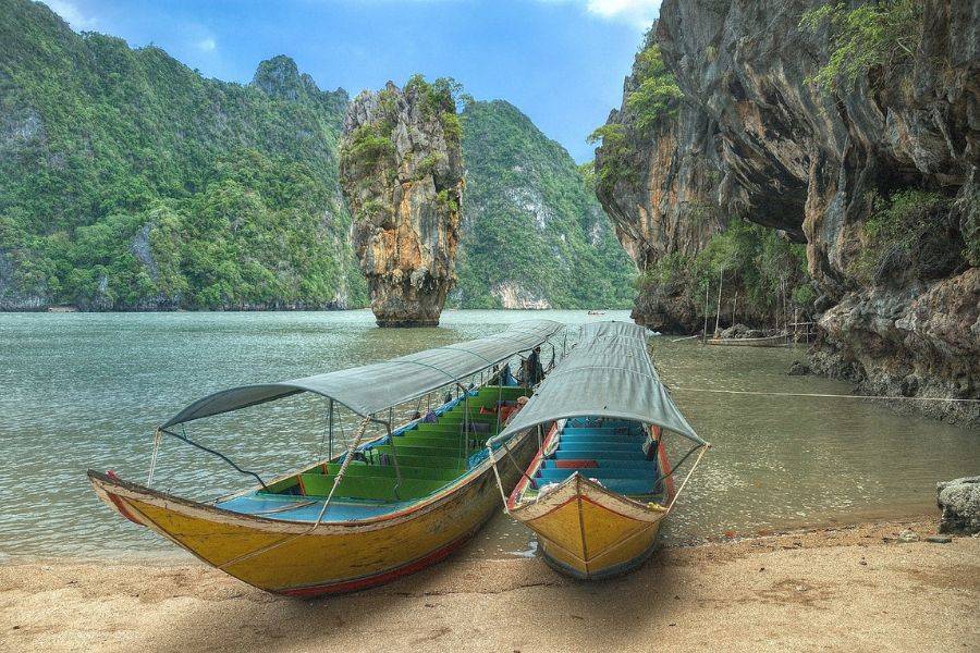 Zwei traditionelle Longtail-Boote mit bunten Dekorationen im klaren, türkisfarbenen Wasser einer Bucht, umgeben von hohen, grünen Kalksteinfelsen unter einem sonnigen Himmel.