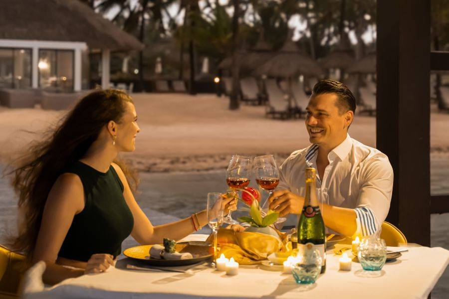  Paar genießt ein romantisches Candle-Light-Dinner am Strand mit Wein, Kerzenlicht und Blick auf das Meer bei Sonnenuntergang.