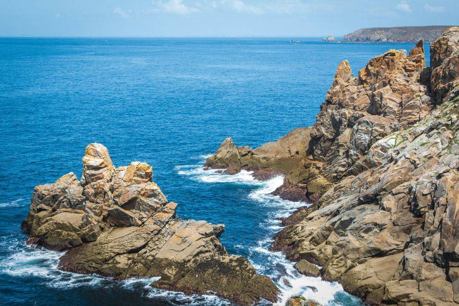 Wuchtige Felsen ragen an der bretonischen Küste aus dem blauen Atlantik, umspült von weißer Gischt unter blauem Himmel.