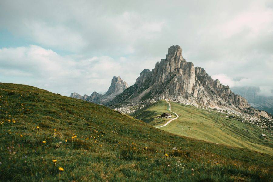 Saftig grüne Wiese mit einem geschwungenen Wanderweg, der zu einem imposanten Felsmassiv in den Dolomiten führt, unter bewölktem Himmel.