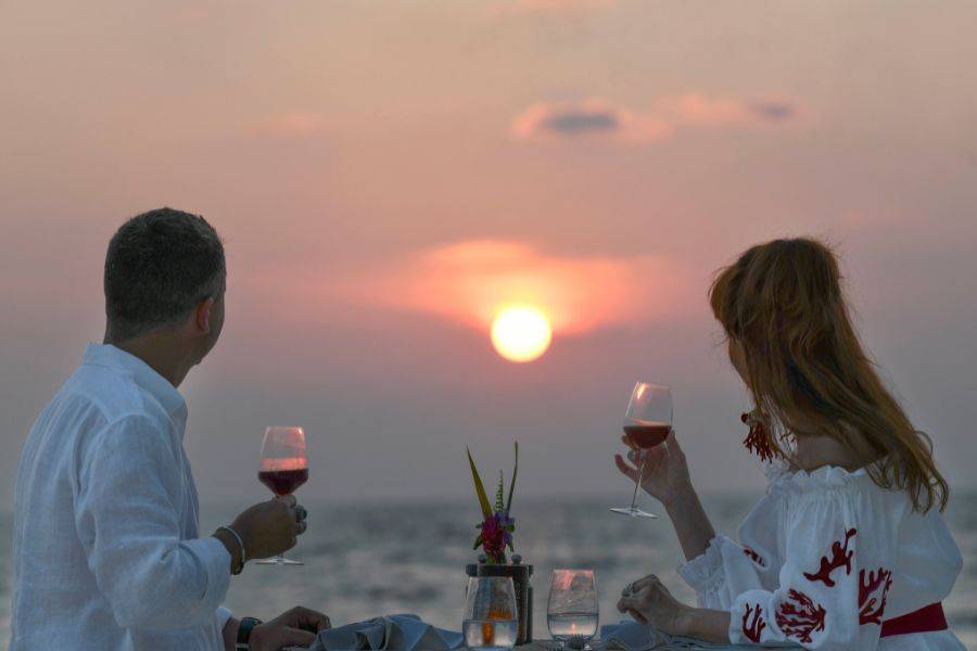 Ein Paar sitzt beim Abendessen und sieht in die Ferne. Die Sonne geht gerade unter und taucht den Himmel in ein orangenes Licht. 