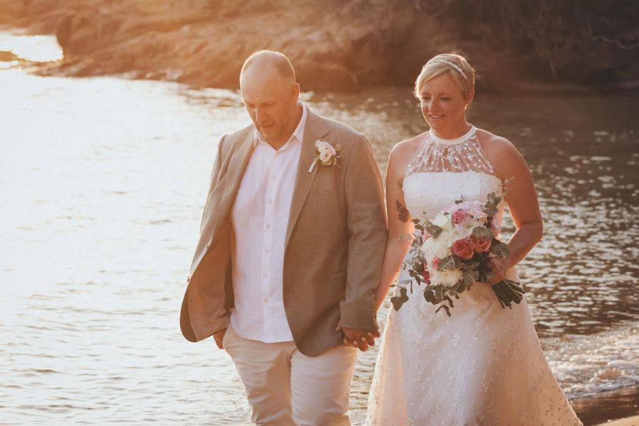 Hochzeitspaar spaziert in der goldenen Abendsonne am Wasser entlang, die Braut trägt einen Blumenschmuck und hält liebevoll den Bräutigam an der Hand.
