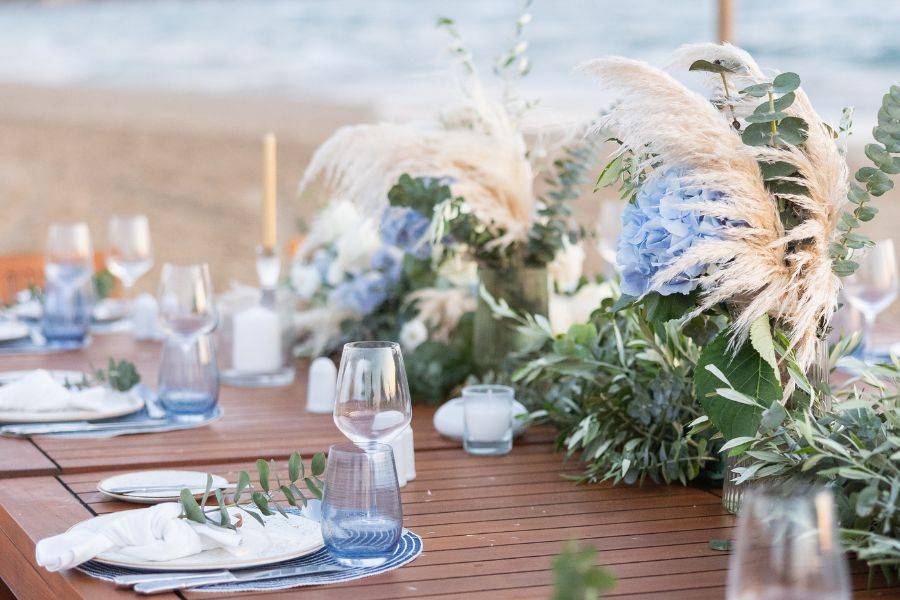 Detailaufnahme der Hochzeitstafel am Strand mit eleganter Blumendekoration aus blauen Hortensien, Pampasgras und grünen Blättern.