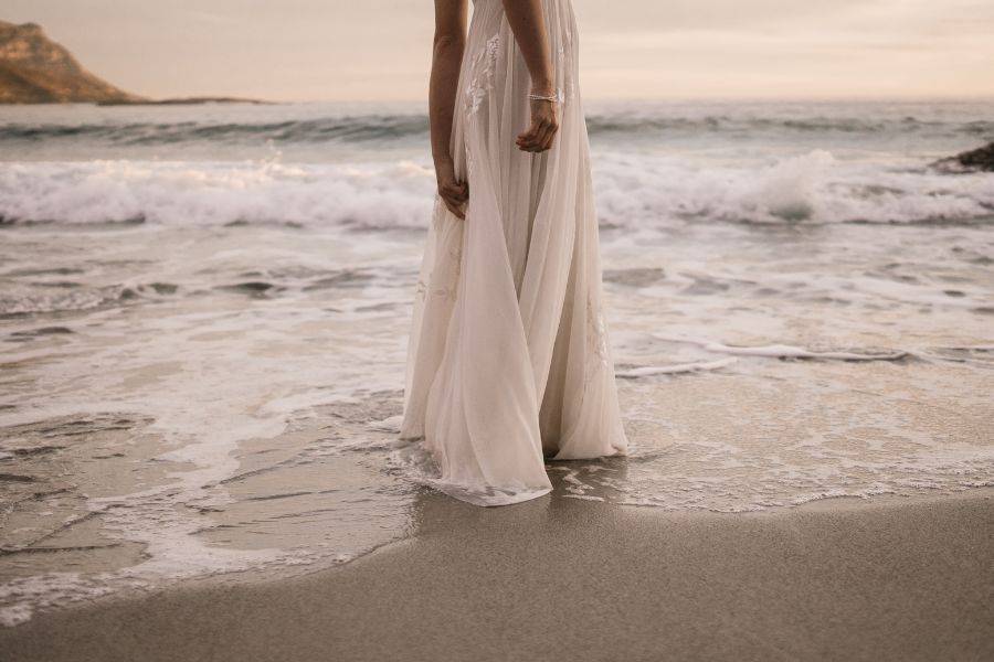 Eine Frau in einem fließenden weißen Kleid steht barfuß am Strand, während die Wellen sanft ihre Füße umspülen. Die Atmosphäre ist romantisch und verträumt.
