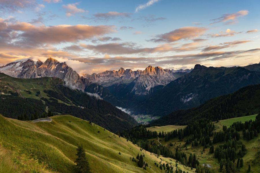 Weite Aussicht auf die Dolomiten bei Sonnenuntergang, mit sanften Hügeln im Vordergrund und dramatisch beleuchteten Berggipfeln im Hintergrund.