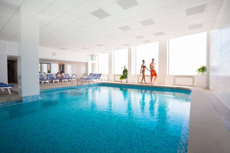 Heller Innenpoolbereich mit blauem Wasser, entspannter Atmosphäre, Liegestühlen, großen Fenstern und Personen, die den Pool genießen.