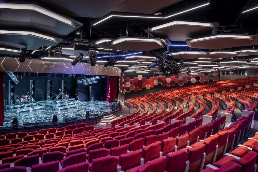 Großes, futuristisches Theater mit roten Sitzen, hexagonalen Deckenpaneelen und einer modern ausgestatteten Bühne mit Scheinwerfern und Technikgerüsten.