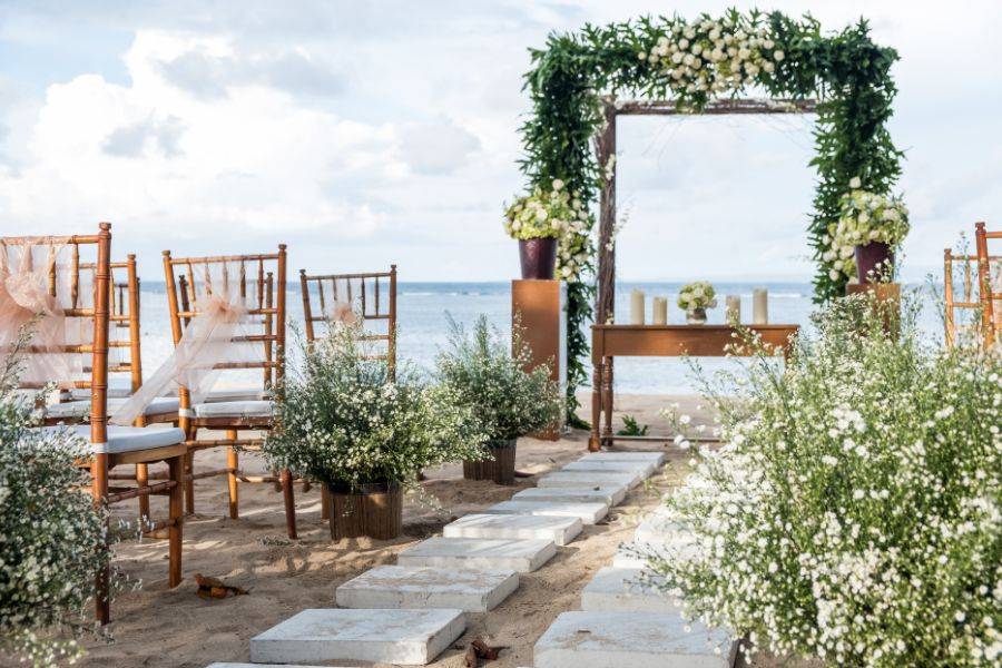 Eine wunderschön dekorierte Hochzeitszeremonie am Strand. Ein mit Blumen geschmückter Bogen steht vor dem Meer, umgeben von Stühlen mit eleganten Verzierungen.