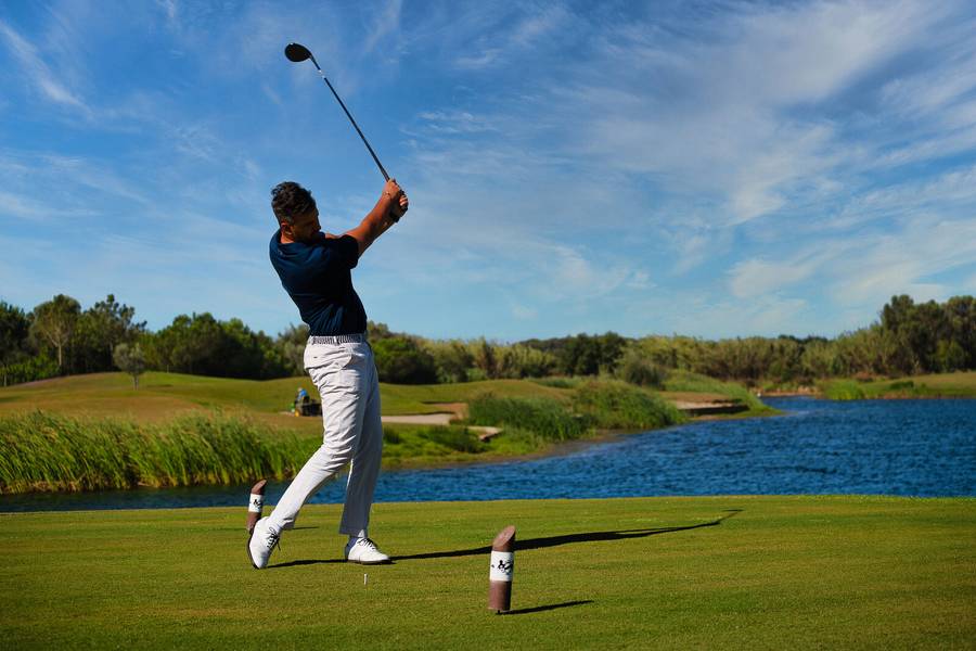Ein Mann in weißer Hose und dunkelblauem Shirt schlägt einen Golfball auf einem gepflegten Golfplatz ab. Im Hintergrund sind ein See, grüne Landschaft und ein blauer Himmel zu sehen.