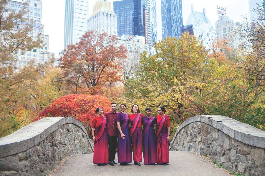 © Air India Air-India-Crew in rot-violetten Uniformen posiert lachend auf einer Brücke im Central Park, mit bunten Herbstbäumen und Wolkenkratzern im Hintergrund.