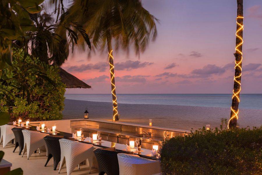 Romantisch beleuchtetes Strandrestaurant mit gedeckten Tischen und Palmen, im Hintergrund ein farbenprächtiger Sonnenuntergang über dem Meer.