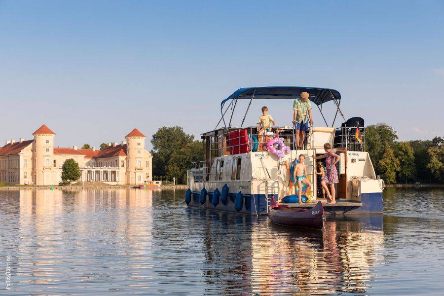 © Ruefa Sicht auf die Hinterseite eines Hausbootes. Man sieht eine Familie mit drei Kindern an Bord. Dahinter sieht man ein Schloss in angenehmer Abendsonne.