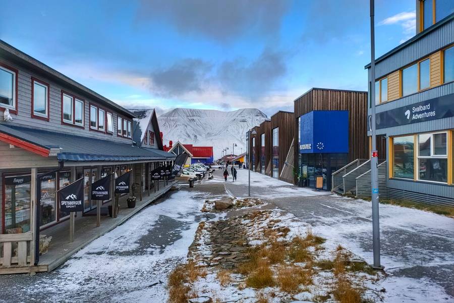 Eine winterliche Straßenansicht mit Gebäuden aus Holz und modernen Fassaden, umgeben von schneebedeckten Bergen im Hintergrund, unter einem klaren Himmel.