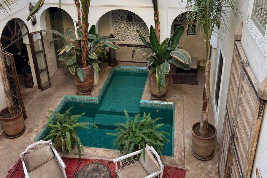 Ein marokkanischer Riad-Innenhof mit einem türkisfarbenen Pool in der Mitte, umgeben von exotischen Pflanzen wie Bananenbäumen und Farnen. Die Architektur ist traditionell mit verzierten Bögen, weißen Wänden und hölzernen Türen im Berberstil.