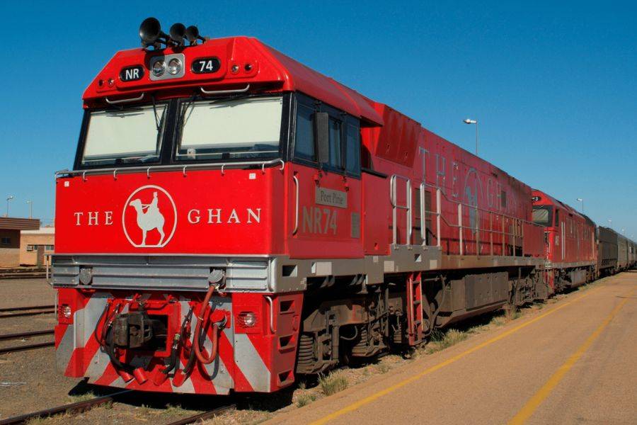 Die rote Lokomotive des berühmten australischen Luxuszuges 'The Ghan' steht auf einem Bahnhofsgleis unter klarem, blauem Himmel. Die Front der Lok trägt das ikonische Logo mit einem Kamel und einem Reiter, das an die historischen afghanischen Kamelkarawanen erinnert.