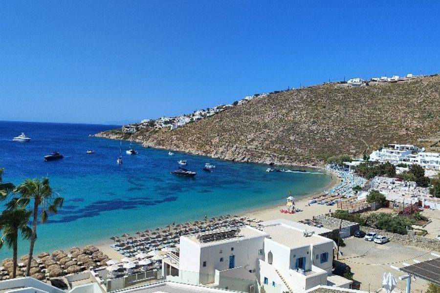 Traumhafte Bucht auf Mykonos mit türkisblauem Wasser, luxuriösen Yachten und weißen Häusern entlang des Hangs – perfekter Blick auf den Strand mit Sonnenliegen und Schirmen unter strahlend blauem Himmel.