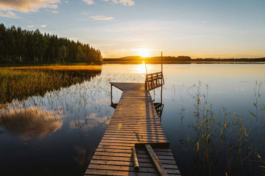 Holzsteg an einem ruhigen See im Norden bei Sonnenuntergang, mit spiegelglattem Wasser, Schilf am Ufer und warmem Abendlicht.