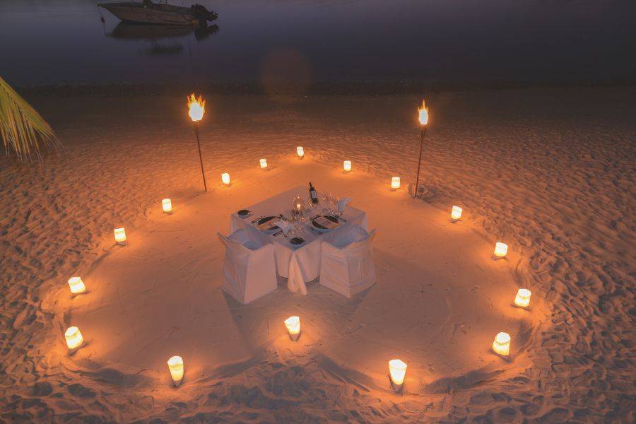 Romantisch gedeckter Tisch am Strand, umgeben von Fackeln und Kerzen im Sand, mit Blick auf ein Boot im ruhigen Wasser bei Nacht.