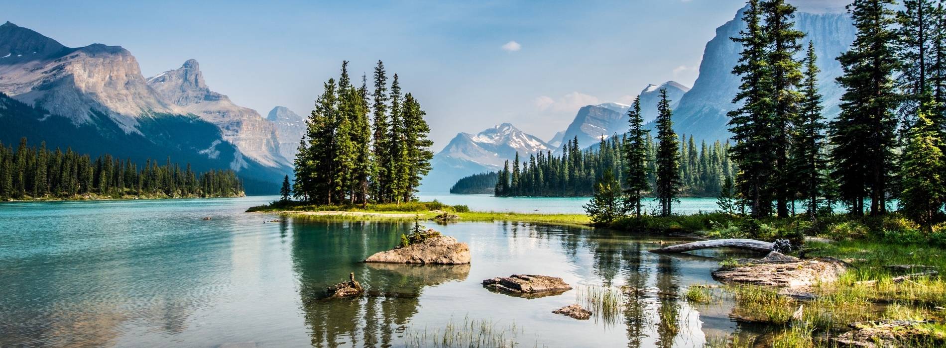 Klarer Bergsee mit kleinen Inseln und Nadelbäumen, umgeben von hohen Bergen unter blauem Himmel