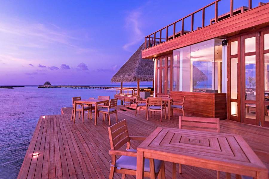 Holzterrasse eines Restaurants am Wasser bei Sonnenuntergang, mit stilvollen Sitzplätzen und Blick auf den ruhigen Ozean.