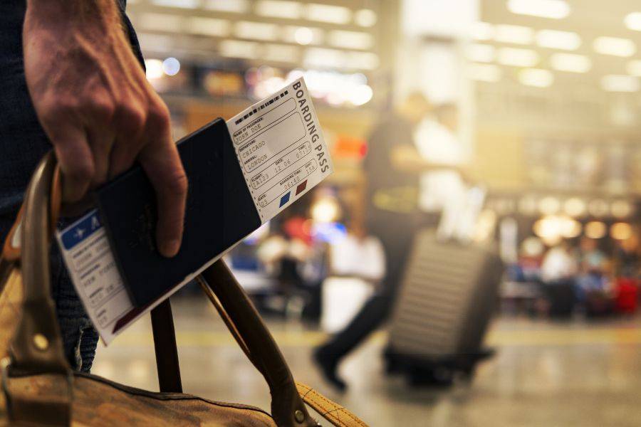 Nahaufnahme einer Hand, die einen Reisepass in der Hand hält. Im Hintergund sieht man einen Flughafen und jemanden, der einen Koffer hinter sich herzieht.