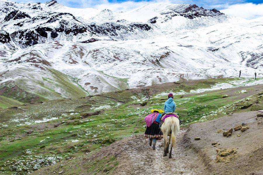 Zwei Menschen in traditioneller Kleidung wandern mit einem Pferd durch eine hochandine Landschaft, teils grün, teils schneebedeckt, mit den schneebedeckten Anden im Hintergrund.