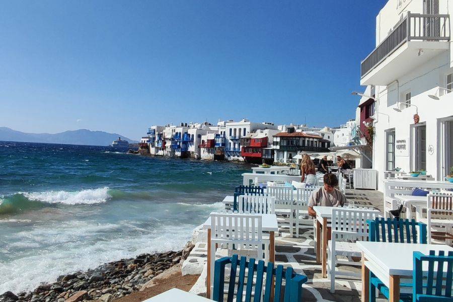 Tisch mit Aussicht – Blick auf das türkisfarbene Meer und die ikonischen weißen Häuser mit blauen und roten Balkonen in Mykonos, Griechenland.
