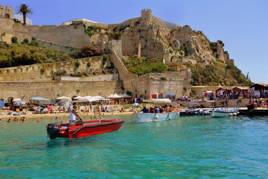 Blick vom Wasser auf den Strand und die historische Festung von Vieste, mit Booten und Badegästen im türkisblauen Meer.