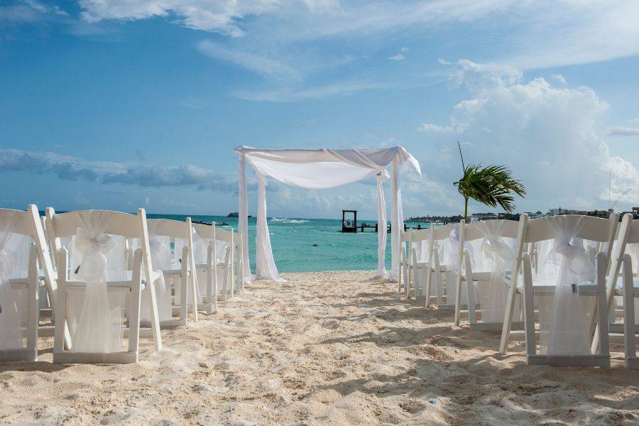  Weiße Hochzeitsstühle und ein dekorierter Traubogen am Sandstrand, türkisfarbenes Meer im Hintergrund – festlich gedeckte Kulisse für eine freie Trauung.