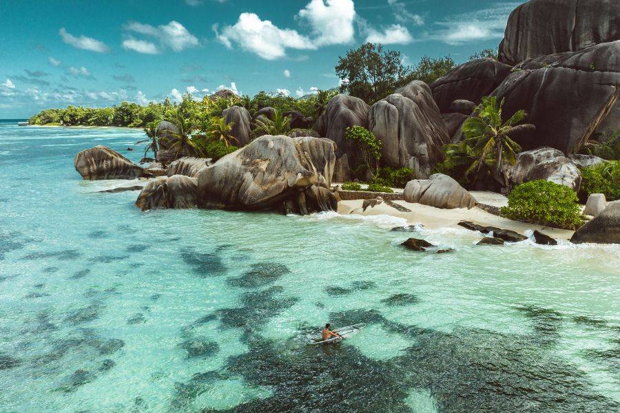 Luftaufnahme eines traumhaften Strandes auf den Seychellen mit türkisblauem Wasser, Granitfelsen, Palmen und einer Person im Wasser – tropische Kulisse ideal für eine romantische Hochzeit oder Flitterwochen.