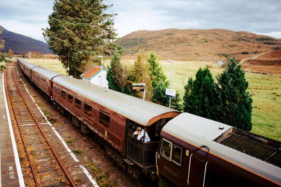 Ein eleganter, dunkelbrauner Luxuszug hält an einem ländlichen Bahnhof in Schottland. Die Zugwaggons erstrecken sich entlang des Bahnsteigs, während ein Mitarbeiter aus einem offenen Fenster lehnt.