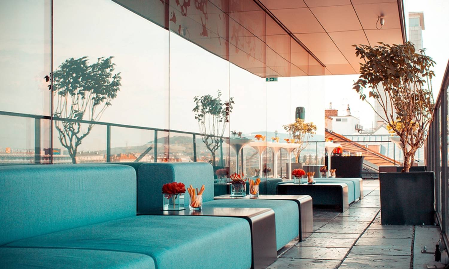 Blick auf eine Dachterasse mit gläserner Front. Davor stehen Lounge Möbel in türkiser Farbe mit Snacks und Tischdeko.
