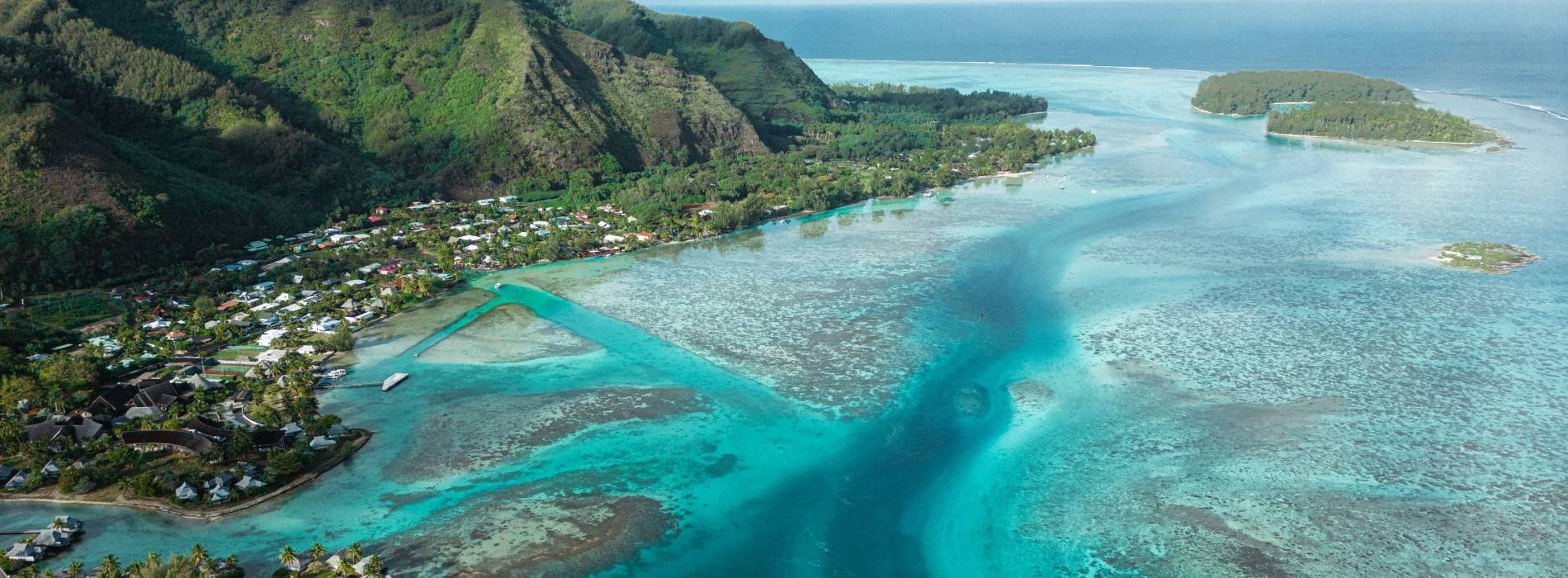 Luftaufnahme einer türkisfarbenen Lagune mit Korallenriffen, Küstenort und grünen Bergen im Hintergrund