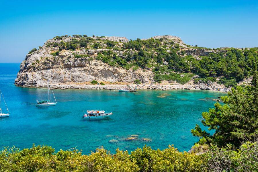 Eine malerische Bucht mit türkisfarbenem Wasser, umgeben von grünen Hügeln und Felsen. Segelboote und kleine Schiffe ankern in der ruhigen Bucht.