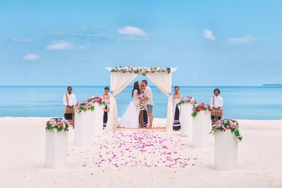 © Sun Siyam Brautpaar küsst sich vor einem blumengeschmückten Pavillon bei einer romantischen Strandhochzeit unter blauem Himmel, umgeben von tropischer Dekoration und Zeremoniengästen.