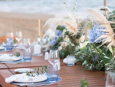 Detailaufnahme der Hochzeitstafel am Strand mit eleganter Blumendekoration aus blauen Hortensien, Pampasgras und grünen Blättern.