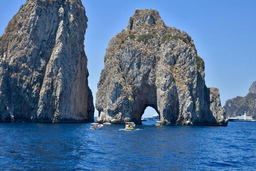 Die markanten Faraglioni-Felsen vor Capri ragen aus dem Meer, mehrere Boote fahren durch das natürliche Felsentor bei strahlendem Sonnenschein.