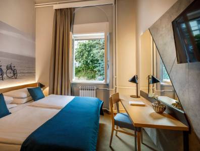 Modernes Hotelzimmer mit zwei Einzelbetten, minimalistischer Einrichtung, sanfter Beleuchtung und einem Fenster mit Blick auf grüne Bäume und ein Gebäude im Hintergrund.