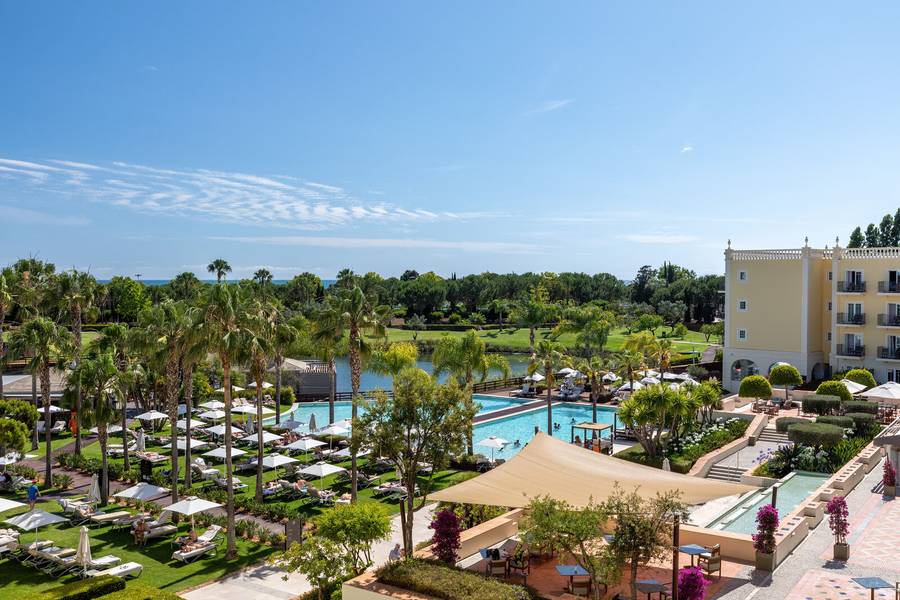 Weitläufige Gartenanlage mit Palmen, Sonnenliegen und mehreren Pools im Domes Lake Algarve. Der Blick reicht über das Resort hinweg bis zum blauen Himmel mit leichten Wolkenformationen.