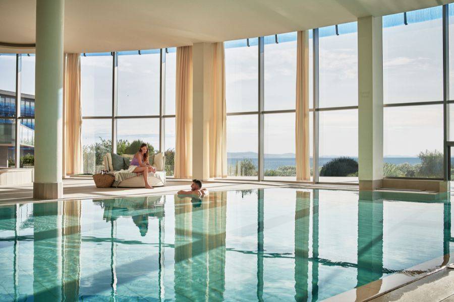 Eleganter Innenpool mit großen Panoramafenstern und Meerblick, eine Person entspannt auf einer Liege im Hintergrund.