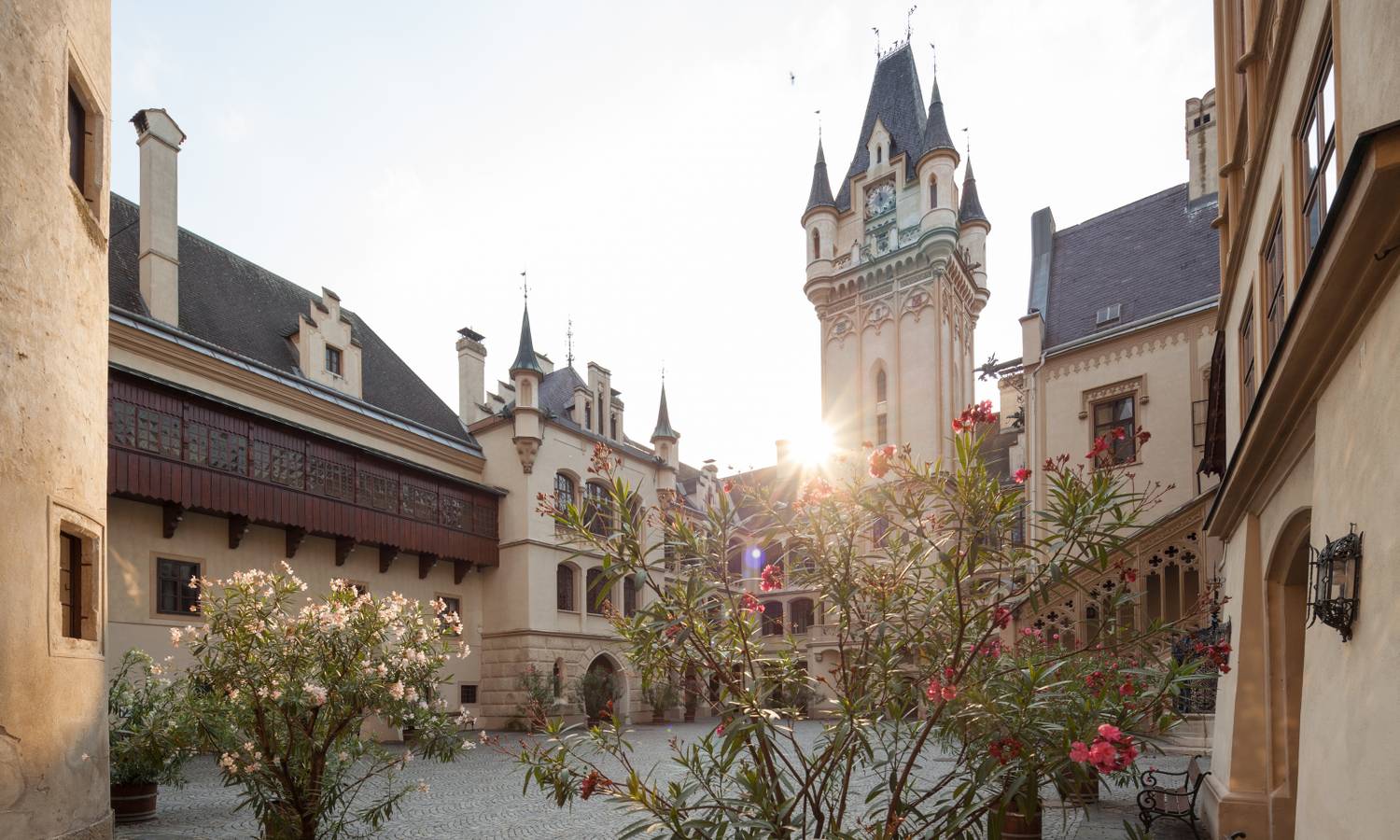 Ein romantischer Schlosshof mit Blumen, Fachwerkbalkonen und einem Turm. Die Sonne scheint durch die Gebäude.