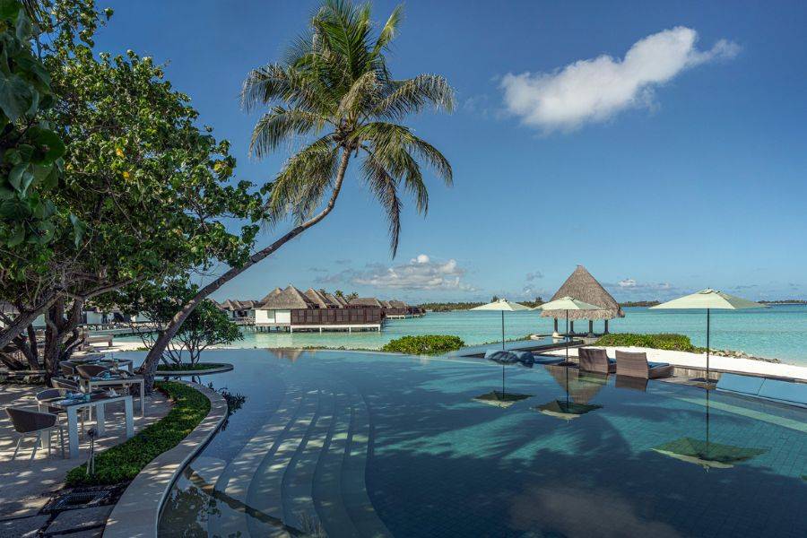 Traumhafter Infinity-Pool mit Blick auf das türkisfarbene Meer, umgeben von Palmen, Sonnenliegen und traditionellen Wasserbungalows im Hintergrund.