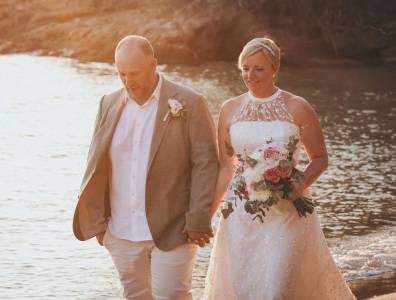 Hochzeitspaar spaziert in der goldenen Abendsonne am Wasser entlang, die Braut trägt einen Blumenschmuck und hält liebevoll den Bräutigam an der Hand.