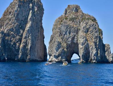 Die markanten Faraglioni-Felsen vor Capri ragen aus dem Meer, mehrere Boote fahren durch das natürliche Felsentor bei strahlendem Sonnenschein.