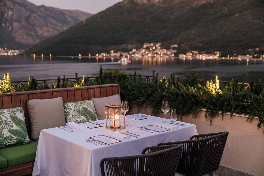 Romantisch gedeckter Tisch auf einer Terrasse mit Blick auf einen ruhigen See und eine malerische Berglandschaft in der Abenddämmerung, beleuchtet von Kerzenlicht und kleinen Lichtern am Ufer.