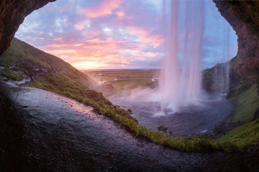 Blick hinter einen Wasserfall in Island bei Sonnenuntergang mit weichem Licht, grüner Landschaft und weiter Ebene im Hintergrund.
